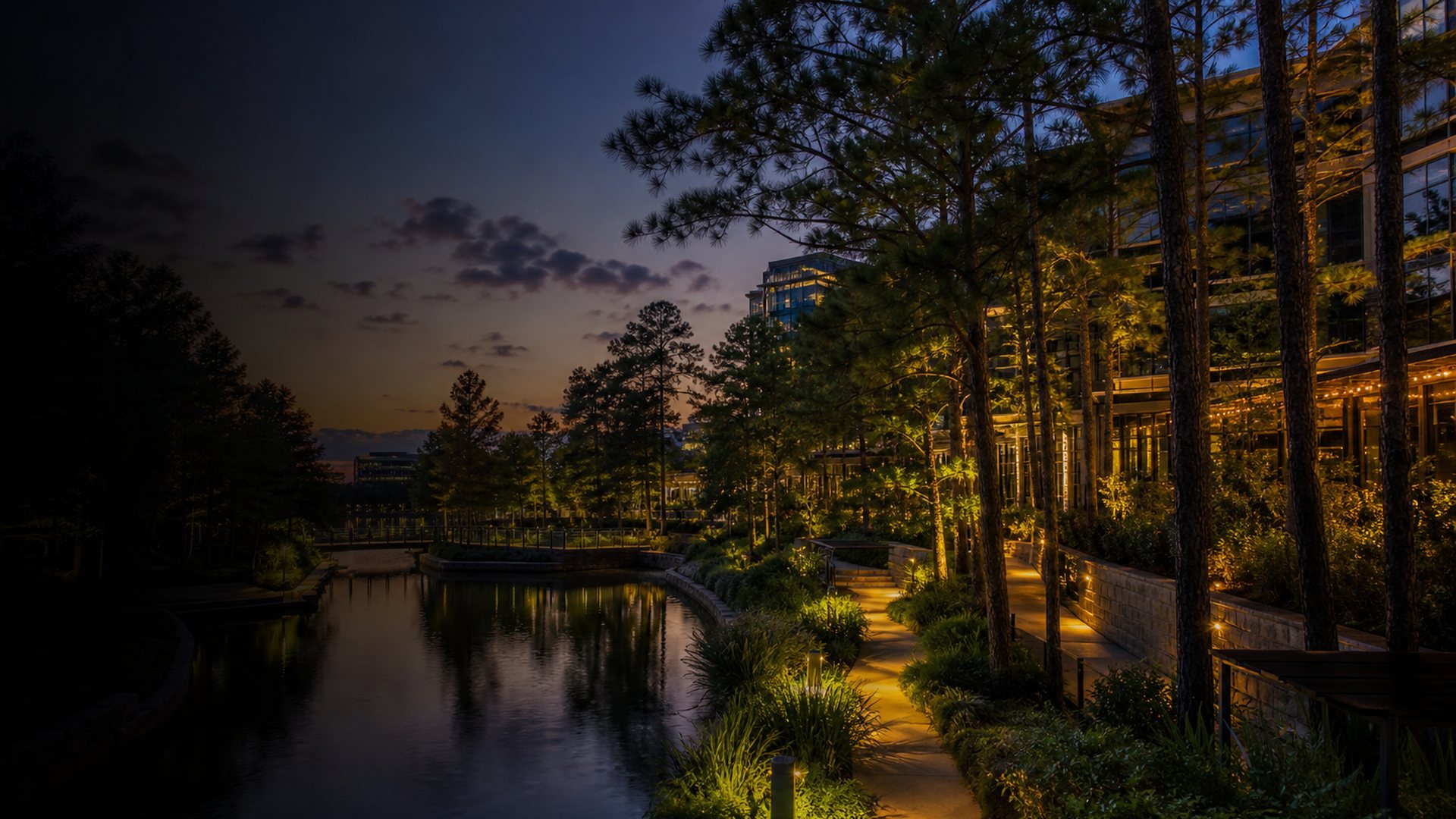 Blue-hour view of The Woodlands, Texas with trees, waterway landscaping, and warm building lights